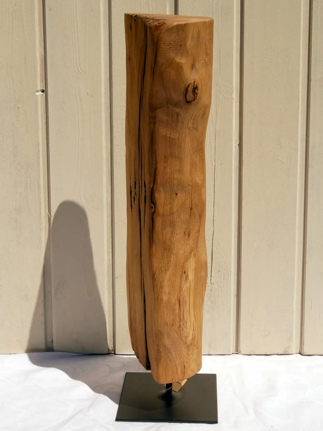 Aufnahme einer rustikalen Eichenholz-Skulptur mit tiefen Maserungen und natürlichen Verwitterungsspuren. Das Holz ist hellbraun und steht fest auf einem flachen, quadratischen, schwarzen Metallsockel. Nachhaltig und handgefertigt.