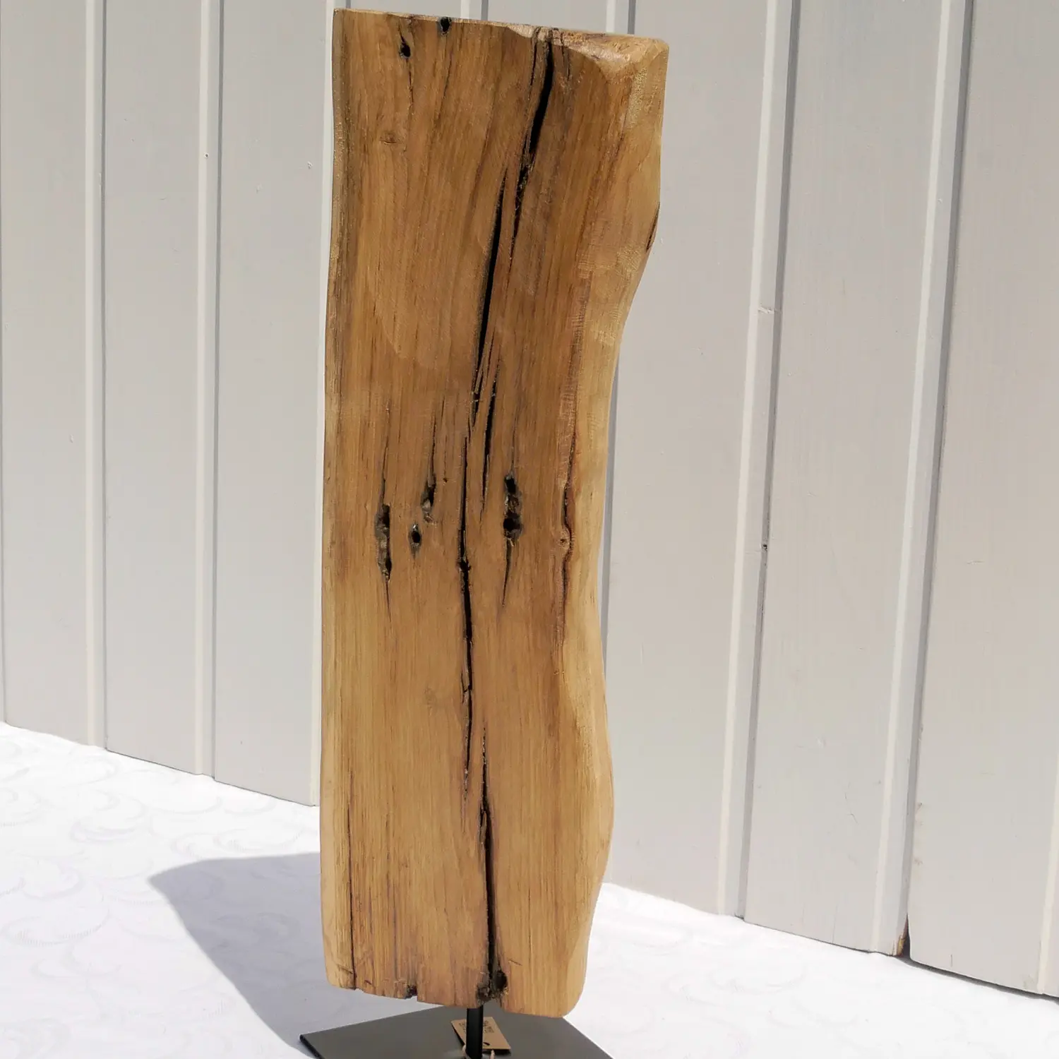 Aufnahme einer rustikalen Eichenholz-Skulptur mit tiefen Maserungen und natürlichen Verwitterungsspuren. Das Holz ist hellbraun und steht fest auf einem flachen, quadratischen, schwarzen Metallsockel. Nachhaltig und handgefertigt.