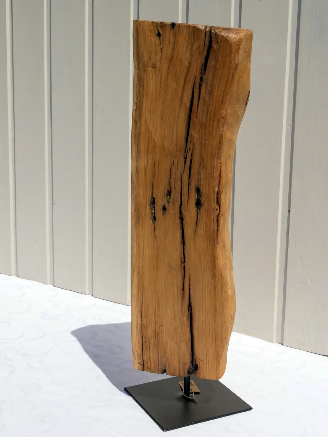 Aufnahme einer rustikalen Eichenholz-Skulptur mit tiefen Maserungen und natürlichen Verwitterungsspuren. Das Holz ist hellbraun und steht fest auf einem flachen, quadratischen, schwarzen Metallsockel. Nachhaltig und handgefertigt.