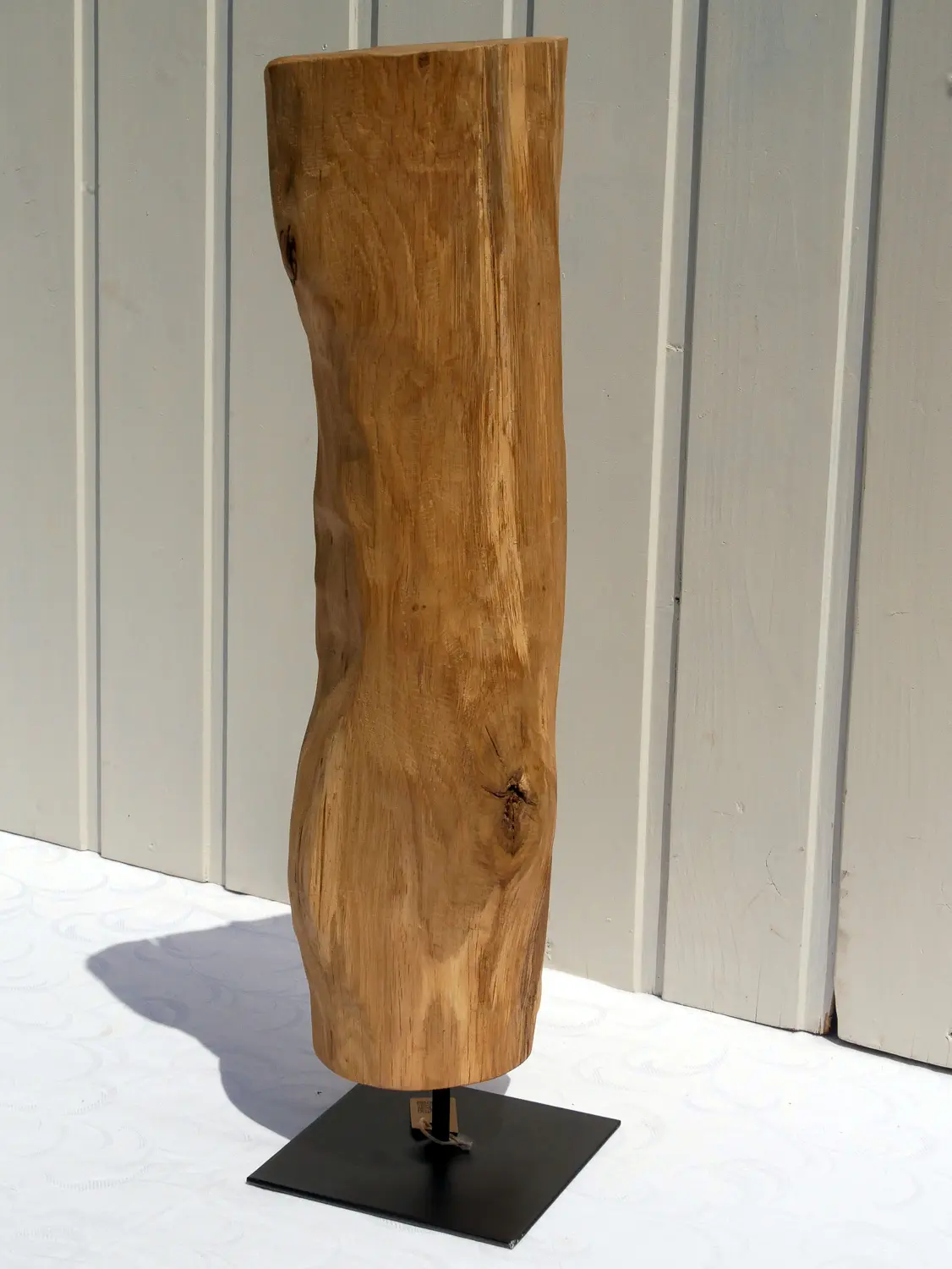Aufnahme einer rustikalen Eichenholz-Skulptur mit tiefen Maserungen und natürlichen Verwitterungsspuren. Das Holz ist hellbraun und steht fest auf einem flachen, quadratischen, schwarzen Metallsockel. Nachhaltig und handgefertigt.
